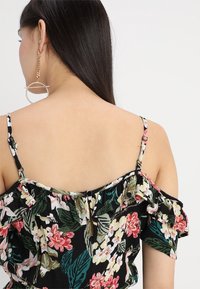 Černý top bez ramínek s květinovým motivem a volánkovými detaily, zdobený růžovými a bílými květy a zelenými listy, s nastavitelnými ramínky a zapínáním na knoflíky.