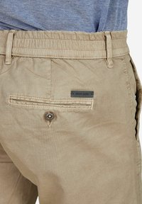 Beige katoenmix broek met een elastische tailleband, achterzak met knoopsluiting en een zwart label met de merknaam.