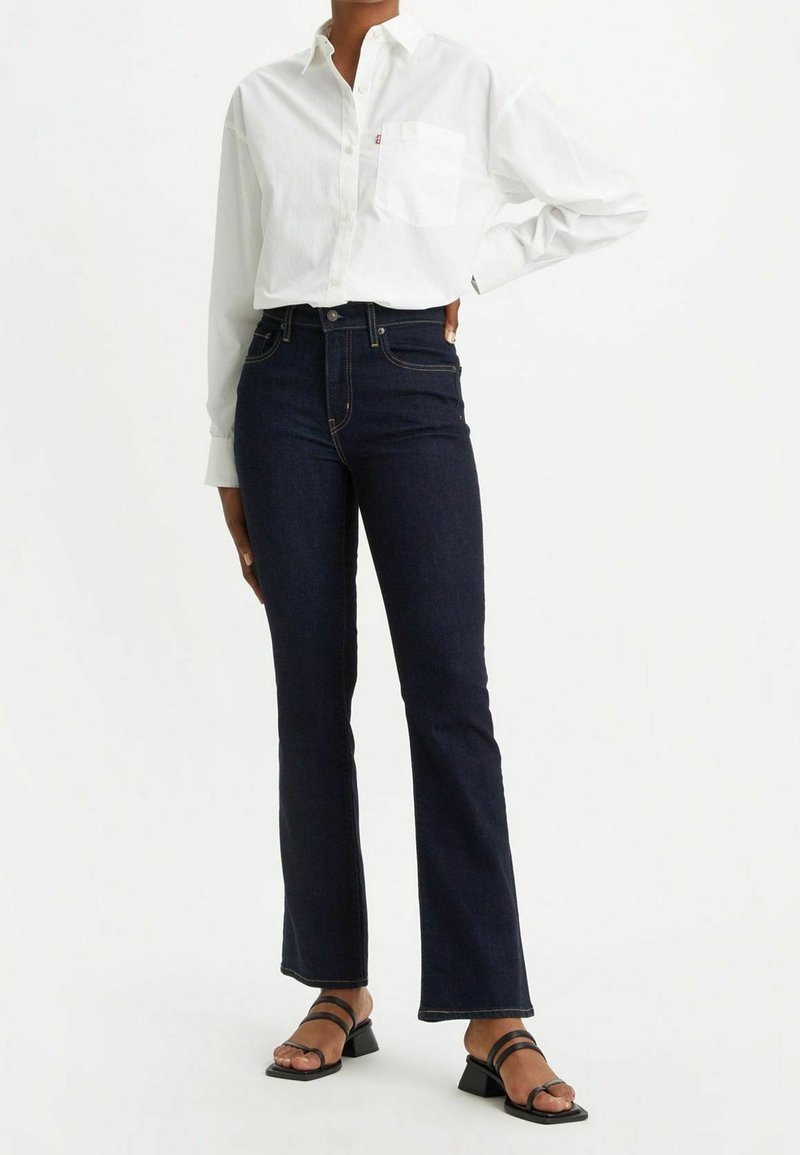 Camisa blanca de botones con bolsillo, jeans de mezclilla oscuros de pata de elefante y sandalias negras de tiras. Textura suave, corte clásico y estilo informal.