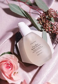 Bottiglia di vetro trasparente con un tappo a pompa bianco, contenente un liquido ambrato chiaro. L'etichetta riporta la dicitura "True Rose Repair Essence." Circondata da un morbido tessuto rosa e fiori.