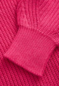 Secțiune de pulover tricotat roz, cu manșete cu zimți, având un model texturat și fir moale, gros. Culoarea vibrantă sporește detaliile vizuale.
