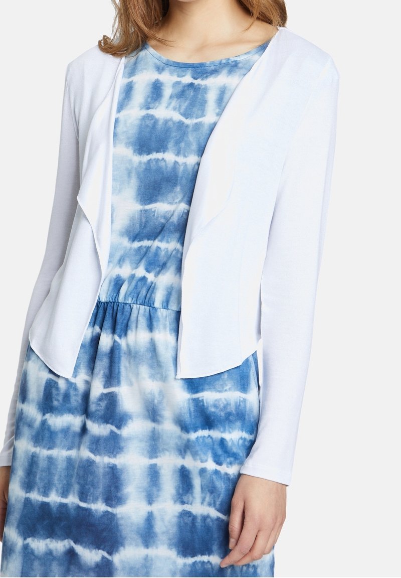 Weiße offene Strickjacke über einem blauen Batik-Kleid. Die Strickjacke hat lange Ärmel und besteht aus einem weichen, leichten Material.