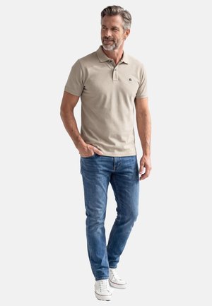 Mann mit grauen Haaren und Bart, trägt ein beiges Poloshirt, blaue Jeans und weiße Sneakers, steht mit einer Hand in der Tasche und schaut nach rechts.