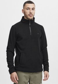 Sudadera de forro polar negro con cuello alto y cierre de media cremallera. Presenta bolsillos laterales y un discreto detalle de logotipo en el pecho. Tejido texturizado.