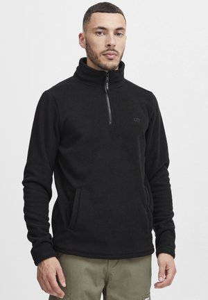 Svart fleece-pullover med høy krage og halv-glidelås. Har sidelommer og subtil logodetalj på brystet. Strukturert stoff.