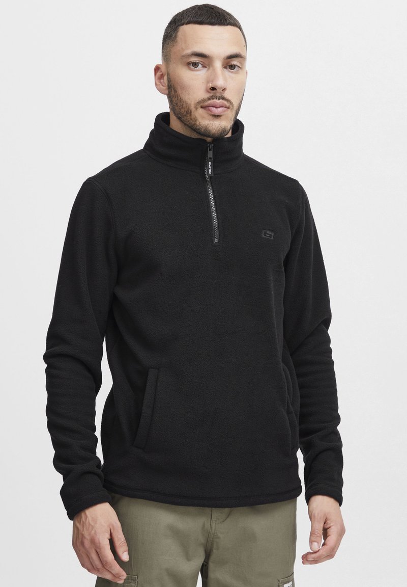 Sudadera de forro polar negro con cuello alto y cierre de media cremallera. Presenta bolsillos laterales y un discreto detalle de logotipo en el pecho. Tejido texturizado.