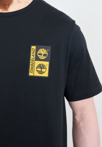 Zwart katoenen t-shirt met een rechthoekig geel en grijs etiket met dubbele boomlogo's, geplaatst op de linkerborst. Korte mouwen.