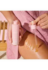Olio snellente in una bottiglia cilindrica rosa con contagocce. L'olio è dorato e brilla sulla pelle, con diverse linee di applicazione visibili.