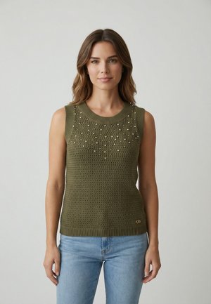 Femme aux cheveux bruns mi-longs portant un haut en tricot sans manches vert olive avec de petites ornements et un jean bleu clair, debout.