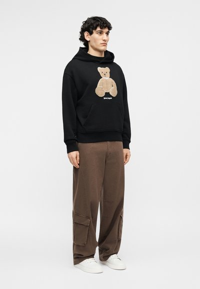 Jonge man met krullend haar, gekleed in een zwarte hoodie met een teddyberenafbeelding, bruine cargobroeken en witte sneakers, staand tegen een effen achtergrond.