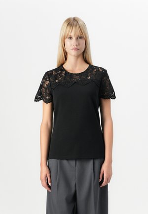 T-shirt noir avec des manches en dentelle courtes à bords festonnés et un col rond, associé à un pantalon large gris.