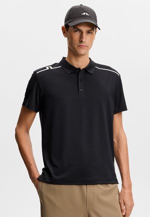 Mand iført sort polotrøje med hvide detaljer på skuldrene, sort kasket med logo og beige bukser, stående mod en hvid baggrund.