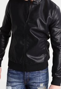 Giacca bomber in pelle nera con colletto tondo, zip frontale e polsini a costine. Abbinata a jeans in denim strappati con dettagli di spruzzi.
