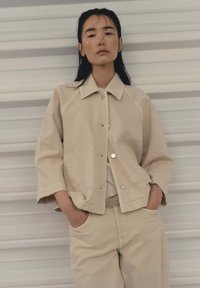 Junge Frau mit langen dunklen Haaren, die eine beigefarbene, geknöpfte Jacke und passende Hosen trägt, steht mit den Händen in den Taschen vor einer gestreiften Wand.