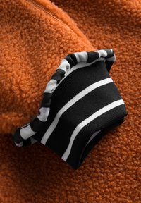 Orange fleece-stof med en tekstureret overflade; sort manchet med hvide striber og en krøllet kantdetalje.