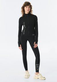Černá sportovní bunda s vysokým límcem a předním zipem, s bílými grafickými akcenty. Spárována s přiléhavými černými legínami. Obě vyrobené z elastického materiálu.