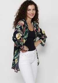 Květinové kimono z tmavého materiálu s zelenými a růžovými tropickými vzory. Lehký design s otevřeným předním dílem a širokými rukávy. Spárováno s bílými džíny.