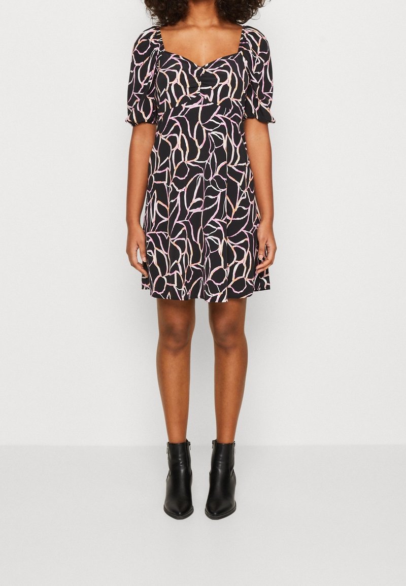 Femme portant une robe courte noire avec des motifs de feuilles roses et beiges, des manches bouffantes et des bottines noires, debout devant un fond clair uni.