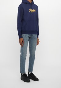 Ung person klädd i en marinblå hoodie med texten "Ralph", ljusa jeans och svarta sneakers, som står mot en enfärgad bakgrund.