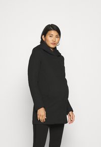 Manteau noir avec un grand col doux, design ajusté, tissu texturé et poches latérales, porté sur des leggings noirs.