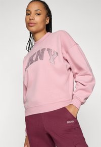 Rózsaszín pulóver kerek nyakú és hosszú ujjú; a DKNY logó csillogó, kontrasztos textúrával díszíti. A modellen összeillő bordó nadrág van.