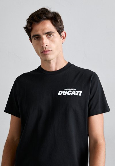 Junger Mann mit kurzen dunklen Haaren, der ein schwarzes T-Shirt mit weißem "DSQUARED2 DUCATI"-Text auf der linken Brust trägt, vor einem schlichten hellen Hintergrund.