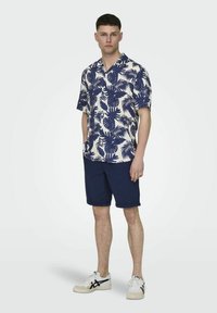 Camicia a maniche corte color navy e crema con motivo a foglie di palma, abbinata a pantaloni corti navy e sneakers bianche con dettagli neri.