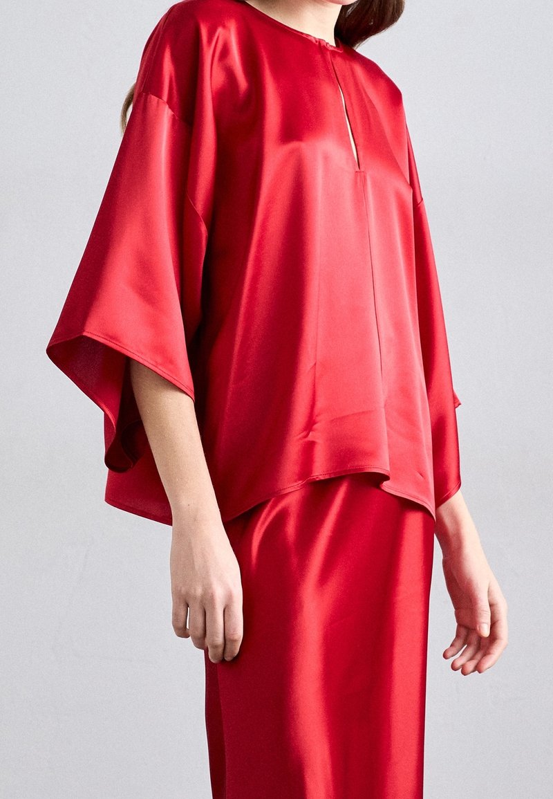 Femme portant un chemisier rouge brillant et ample avec des manches larges et une jupe assortie, montrée des épaules aux hanches sur fond uni.