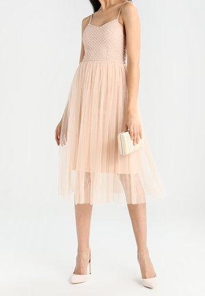 Robe sans manches rose clair avec un corsage texturé et une jupe en tulle évasée. Le mannequin tient un petit sac à main crème et porte des talons nus.