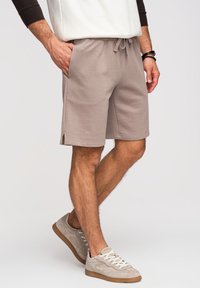 Homme portant un short beige à cordon et des baskets en daim gris clair, debout avec une main dans la poche sur fond blanc.