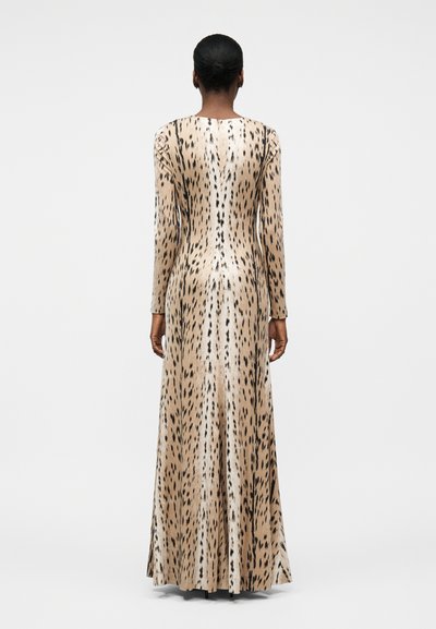 Γυναίκα με κοντά μαλλιά που φοράει μακρύ, εφαρμοστό μπεζ φόρεμα με ζωικό σχέδιο, όρθια με την πλάτη γυρισμένη προς ένα απλό λευκό φόντο.