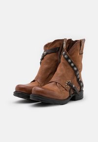 Stivaletti alla caviglia in pelle marrone con una texture leggermente invecchiata, dotati di una cerniera laterale, borchie decorative e una forma curvata.