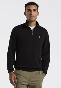 Schwarzer Zip-Pullover aus glattem Stoff. Mit gerippten Bündchen und einem kleinen Logo auf der linken Brust. Kombiniert mit khakifarbigen Hosen.