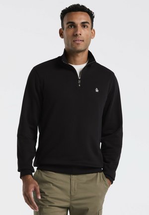 Schwarzer Zip-Pullover aus glattem Stoff. Mit gerippten Bündchen und einem kleinen Logo auf der linken Brust. Kombiniert mit khakifarbigen Hosen.