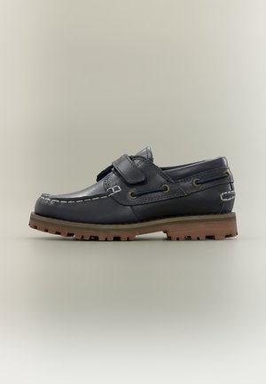 Pantof de barcă din piele albastru închis cu cusături albe, curea cu velcro, talpă robustă maro și detaliu de șireturi bleumarin pe un fundal neutru.