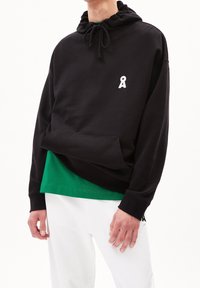 Felpa nera con cappuccio e coulisse, tasca frontale e logo in nastro bianco. Indossata sopra una maglietta verde, abbinata a pantaloni bianchi.