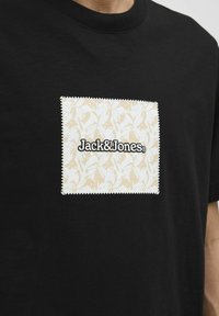 Sort bomulds T-shirt med en tekstureret, cremefarvet firkantet lap med et blomstermønster og "Jack&Jones" logo i sort.
