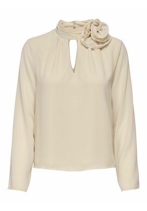 Blouse crème à manches longues avec col montant, découpe en forme de trou de serrure et grande fleur en tissu sur l'épaule gauche.