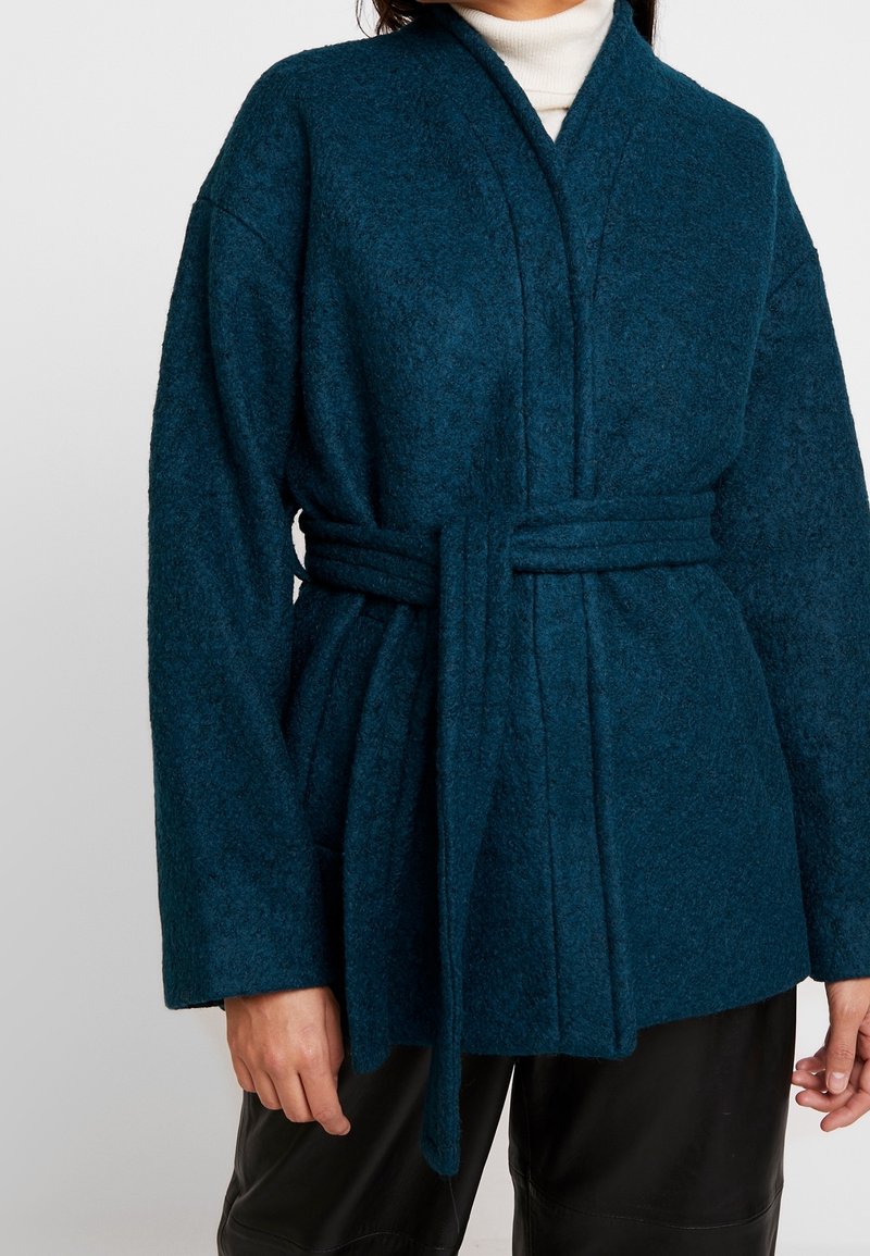 Femme portant un manteau ceinturé texturé couleur bleu sarcelle sur un col roulé blanc et un pantalon noir, posant devant un fond uni.