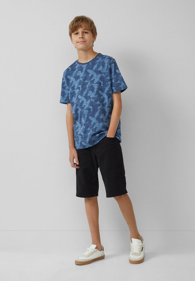 Blauw t-shirt met korte mouwen en abstract patroon, gecombineerd met zwarte shorts. Model draagt witte sneakers met een tan zool. Schoon, minimalistisch ontwerp.