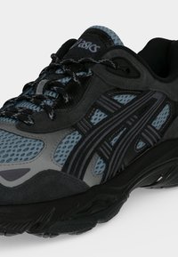 Μαύρο και μπλε παπούτσι για τρέξιμο Asics με υφασμάτινο πλέγμα με σχέδιο, παχιά μαύρη σόλα και μαύρα κορδόνια με γκρι λεπτομέρειες.