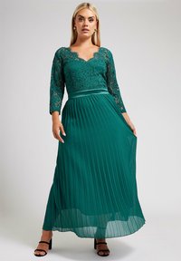 Lång grön plisserad klänning med en spetsliv, med en scalloped halsringning och trekvartsärmar. Accenterad med ett satinmidjeband.