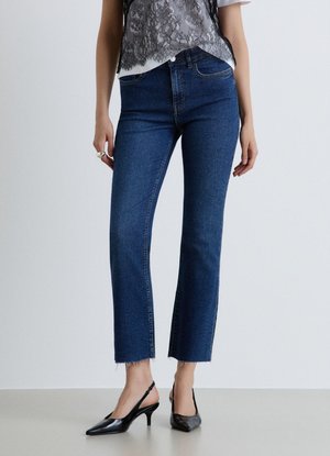 Persona che indossa jeans blu scuro a gamba dritta con orli raw, una maglietta bianca con dettagli in pizzo nero e décolleté slingback nere.