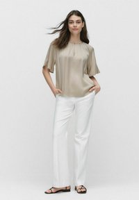 Beige, kortärmad sidenblus med en centralt veck, tillsammans med vita raka byxor och svarta platta sandaler.