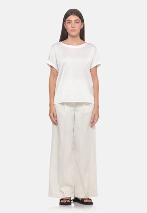Donna con capelli castani lisci che indossa una camicetta bianca a maniche corte, pantaloni bianchi a gamba larga e sandali neri, in piedi su sfondo bianco.