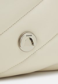 Romige leren handtas met een diamantvormig quiltontwerp, voorzien van een ronde metalen sluiting gegraveerd met "PINKO" en contrasterende stikseldetails.