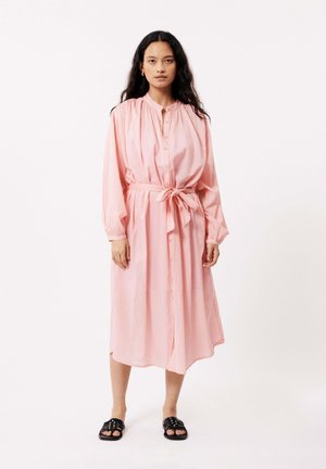 Roze, lang mouw, knopenjurk met een geplooide halslijn en een strikceintuur in de taille. Model draagt zwarte sandalen.