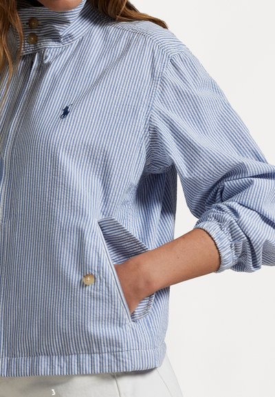 Jaqueta listrada em azul claro e branco com gola alta, punhos elásticos e bolsos laterais. Apresenta um pequeno logótipo perto do peito.
