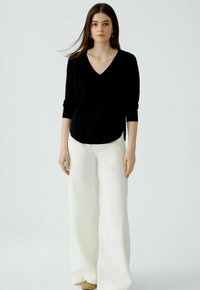 Schwarzer, langärmeliger V-Ausschnitt-Pullover kombiniert mit locker sitzenden weißen Hosen. Neutrale Schuhe, minimaler Schmuck. Glatte Texturen, schlichtes Design.