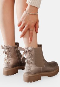 Bottines en suède marron avec une finition lisse, fermeture éclair latérale et cordon décoratif en ruban avec des accents en strass à l'arrière. Semelle épaisse.